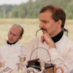 Two men in historical attire sit at a table outdoors, engaging in a reenactment.