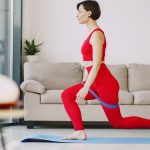 Side view of fit female in sportswear with short hair doing exercise with elastic band on blue mat near sofa in living room